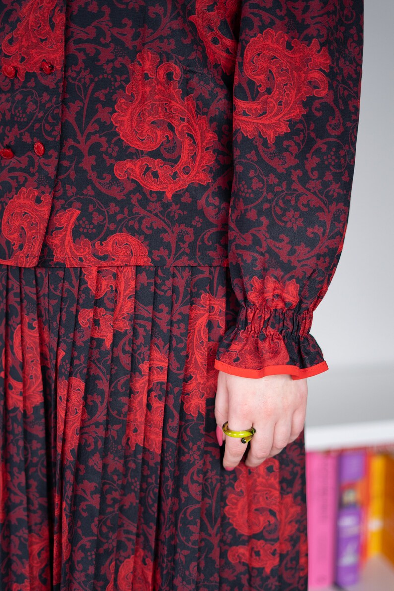 K&ouml;nnte beinhalten: Ein schwarzes und rotes Kleid mit Blumenmuster und einem Faltenrock. Das Kleid hat lange &Auml;rmel mit einer R&uuml;schenmanschette. Das Kleid wird von einer Person mit einem gr&uuml;nen Ring am Finger getragen.