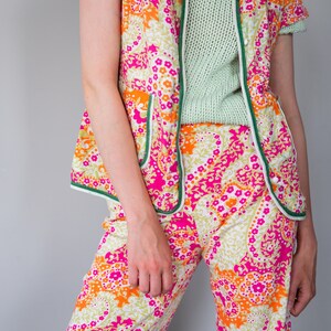May include: A pink and orange floral print pajama set with green trim. The top is a short-sleeved jacket with a button closure. The pants are wide-legged and have a relaxed fit.