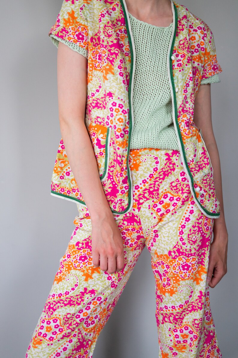 May include: A pink and orange floral print pajama set with green trim. The top is a short-sleeved jacket with a button closure. The pants are wide-legged and have a relaxed fit.