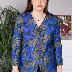 May include: A woman wearing a blue and green floral patterned button-up blouse with gold buttons. She is wearing black pants.