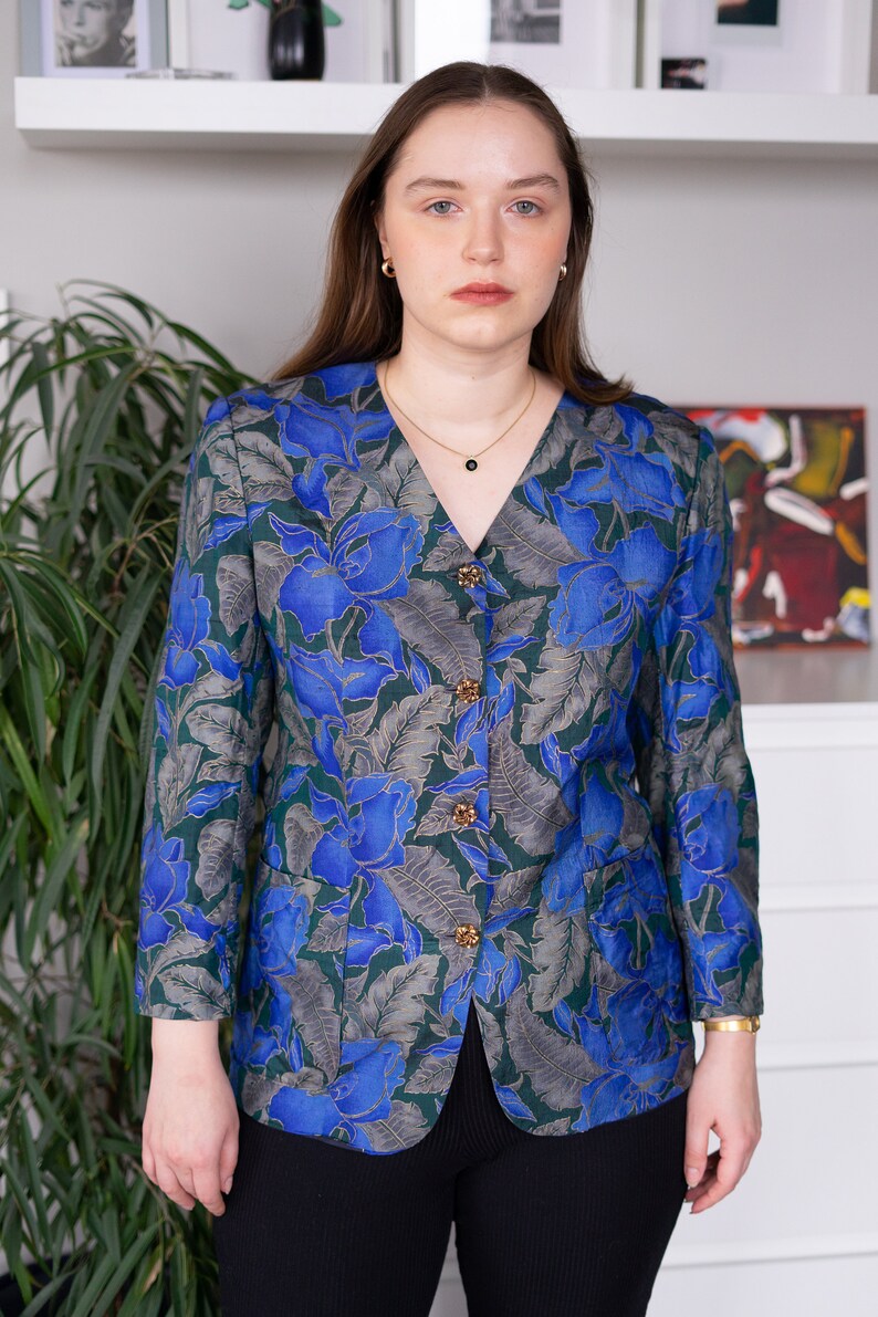 May include: A woman wearing a blue and green floral patterned button-up blouse with gold buttons. She is wearing black pants.