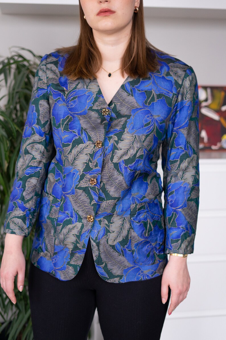 May include: A blue and green floral patterned blazer with gold buttons. The blazer has a v-neck and long sleeves.