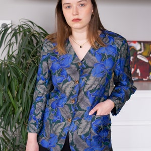 May include: A woman wearing a blue and green floral patterned button-up shirt with gold buttons. She is wearing black pants and has her hand in her pocket.