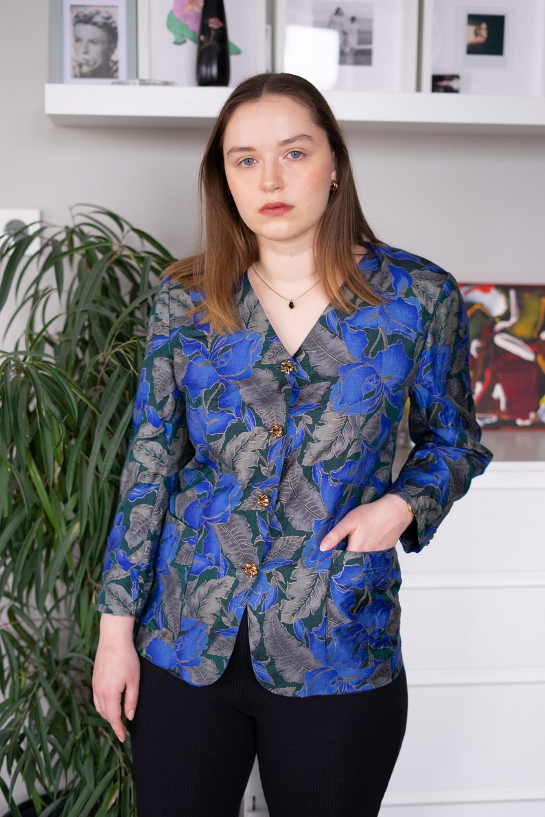 May include: A woman wearing a blue and green floral patterned button-up shirt with gold buttons. She is wearing black pants and has her hand in her pocket.