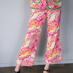 May include: A woman is wearing a pink, orange, and green patterned pantsuit. The pants are wide-legged and the top has a collar and buttons. She is wearing black heeled sandals.