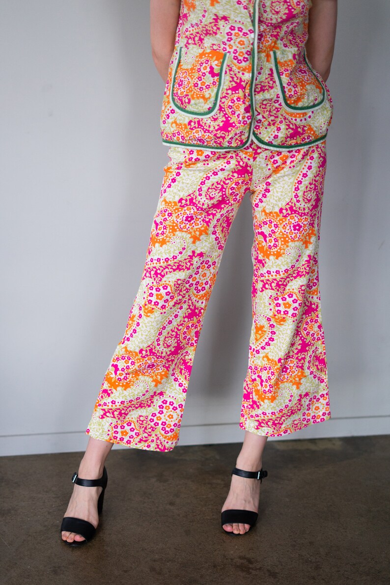 May include: A woman is wearing a pink, orange, and green patterned pantsuit. The pants are wide-legged and the top has a collar and buttons. She is wearing black heeled sandals.