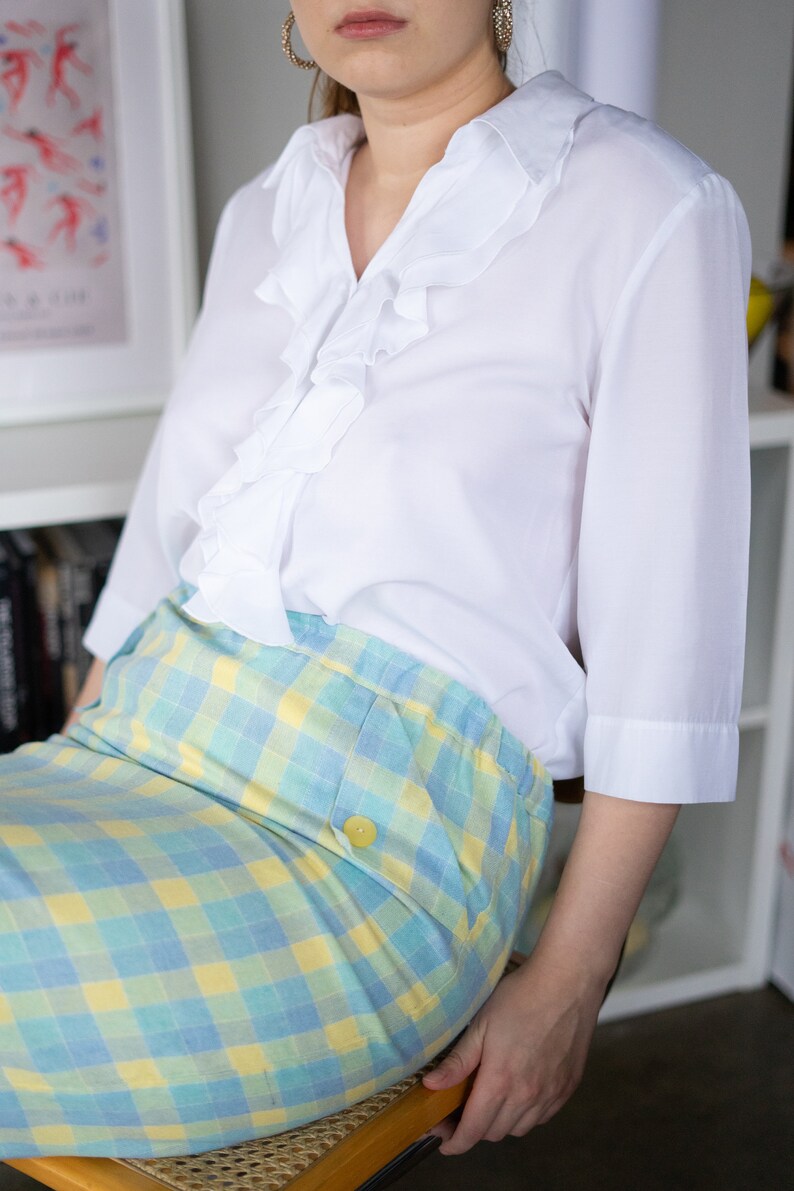 May include: A white blouse with a ruffled collar and a blue and yellow checked skirt.