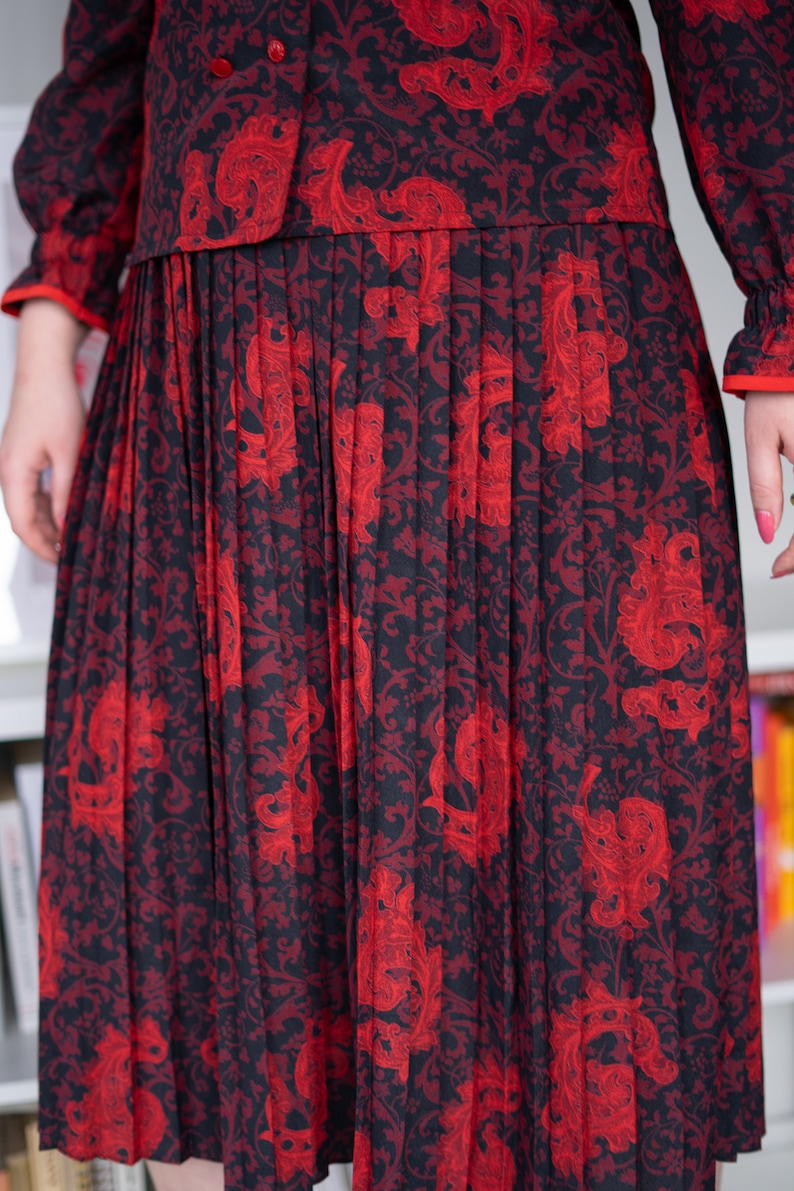 K&ouml;nnte beinhalten: Ein schwarzer und roter Plisseerock mit einem Blumenmuster. Der Rock ist aus einem weichen, flie&szlig;enden Stoff gefertigt und hat ein klassisches, zeitloses Design.