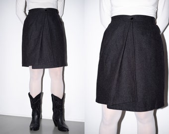 May include: A dark gray wrap skirt with a single button closure at the waist. The skirt is made of a textured fabric and has a slightly A-line shape.