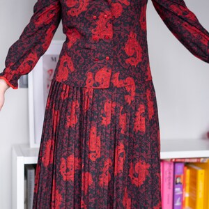 K&ouml;nnte beinhalten: Ein rot-schwarzes gemustertes Kleid mit einem Faltenrock und einem wei&szlig;en Kragen mit rotem Besatz. Das Kleid hat einen Knopfleisten vorne und lange &Auml;rmel.