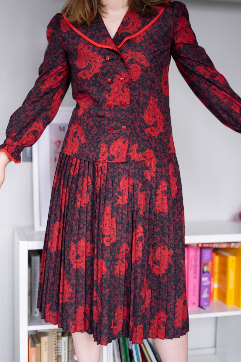 K&ouml;nnte beinhalten: Ein rot-schwarzes gemustertes Kleid mit einem Faltenrock und einem wei&szlig;en Kragen mit rotem Besatz. Das Kleid hat einen Knopfleisten vorne und lange &Auml;rmel.