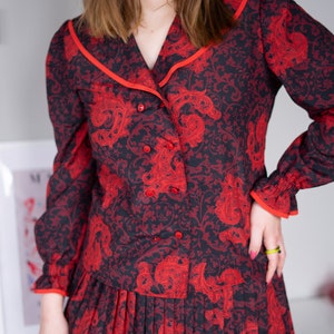 K&ouml;nnte beinhalten: Ein rotes und schwarzes Kleid mit Paisley-Muster, einem Kragen-Oberteil und einem Faltenrock.