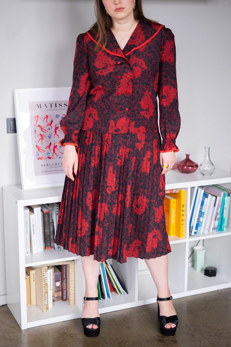 K&ouml;nnte beinhalten: Ein rotes und schwarzes Blumenkleid mit einem Faltenrock und einem Kragenoberteil. Das Kleid hat einen roten Besatz um den Kragen und die &Auml;rmel. Das Model tr&auml;gt schwarze Plateau-Heels.