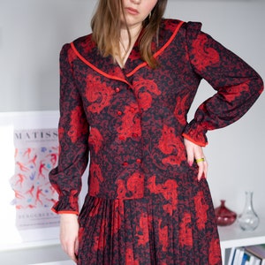 K&ouml;nnte beinhalten: Ein rot-schwarzes gemustertes Kleid mit wei&szlig;em Kragen und rotem Besatz. Das Kleid hat einen Knopfleisten vorne und einen Faltenrock.
