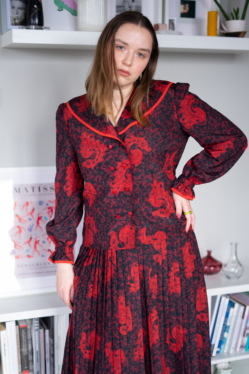 K&ouml;nnte beinhalten: Ein rot-schwarzes gemustertes Kleid mit wei&szlig;em Kragen und rotem Besatz. Das Kleid hat einen Knopfleisten vorne und einen Faltenrock.