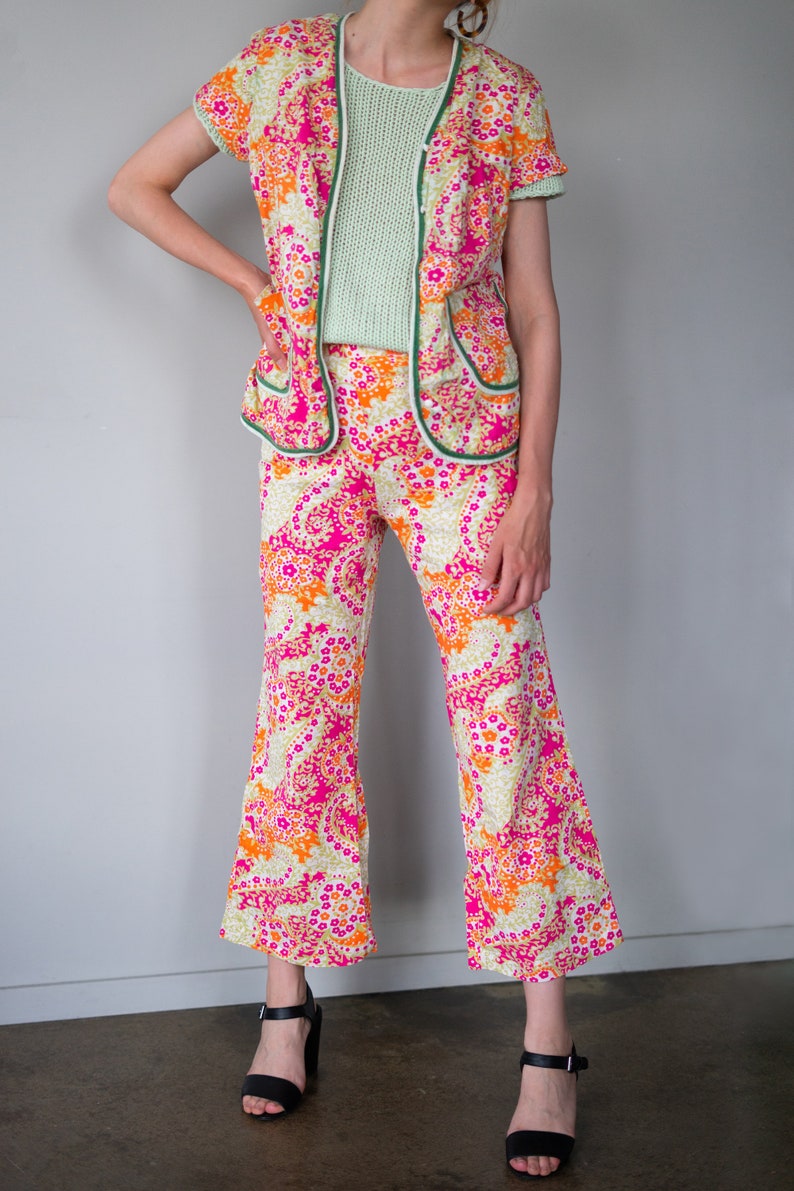 May include: A pink, orange, and white floral print pantsuit with a green trim. The pants are flared and the jacket has a collar and short sleeves.