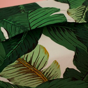 May include: Close-up of a white fabric with a green and brown tropical leaf pattern. The fabric has a seam running along the top edge.