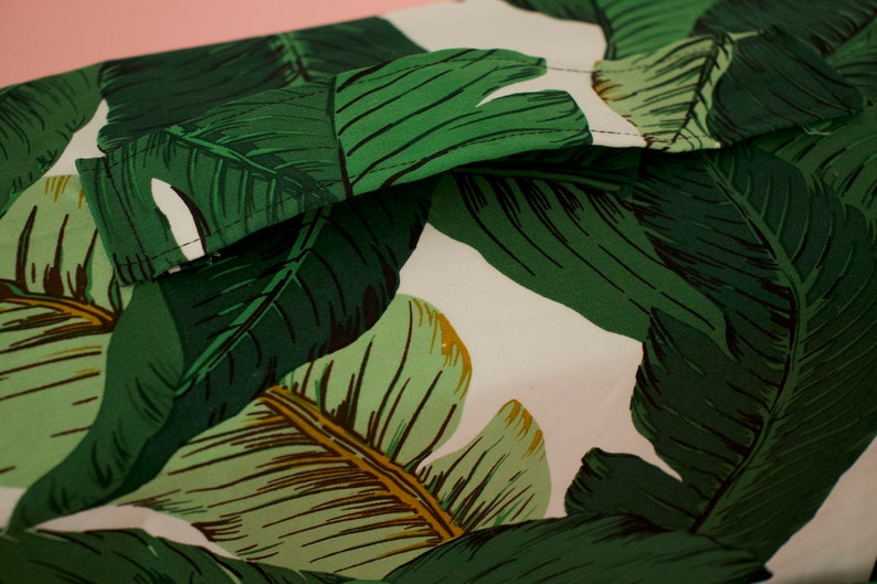 May include: Close-up of a white fabric with a green and brown tropical leaf pattern. The fabric has a seam running along the top edge.