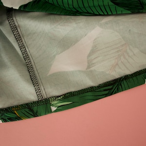 May include: Close-up of the inside seam of a green and white tropical print fabric. The fabric has a white background with green leaves and a black stitching detail.