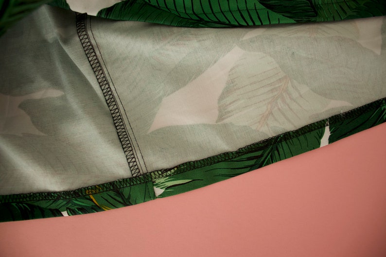 May include: Close-up of the inside seam of a green and white tropical print fabric. The fabric has a white background with green leaves and a black stitching detail.