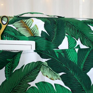 May include: A white sewing machine cover with a green and white banana leaf pattern. The cover has a pocket on the front with a pair of gold scissors and a book titled "BEDIENUNGSANLEITUNG" inside.