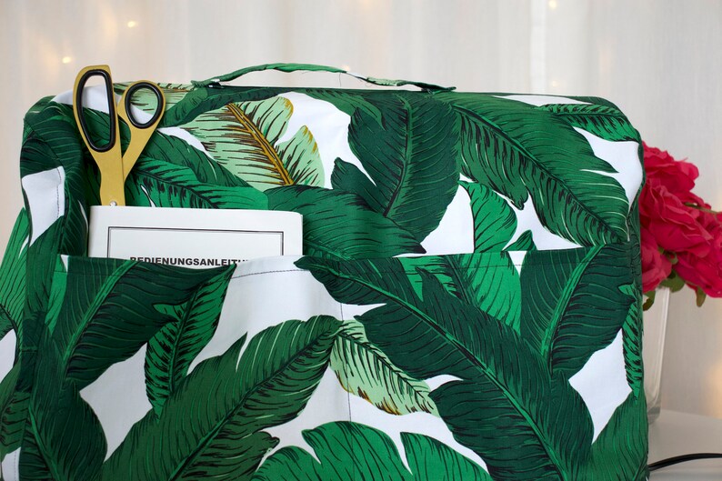 May include: A white sewing machine cover with a green and white banana leaf pattern. The cover has a pocket on the front with a pair of gold scissors and a book titled "BEDIENUNGSANLEITUNG" inside.