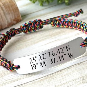 May include: A colorful braided bracelet with a silver metal tag engraved with the coordinates "25° 22' 76.42" N 19° 44' 32.14" W".