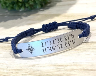 May include: A navy blue braided bracelet with a silver tag engraved with the coordinates "23°32'30.81"N 11°46'52.05"W" and a compass rose.