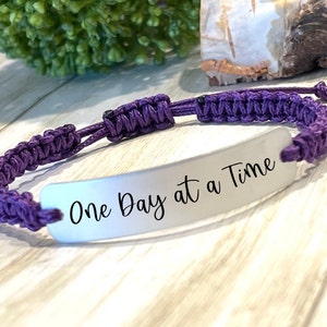 May include: Purple braided cord bracelet with a silver charm that reads "One Day at a Time".