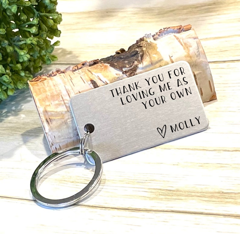May include: A silver rectangular keychain with the text "THANK YOU FOR LOVING ME AS YOUR OWN" and the name "MOLLY" below a heart symbol. The keychain is attached to a silver key ring. The background includes a piece of wood and greenery.