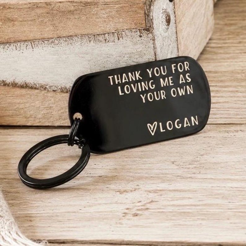 May include: A black metal keychain with a rounded rectangular tag. The tag has the text "THANK YOU FOR LOVING ME AS YOUR OWN" and the name "LOGAN" below, with a heart symbol. The keychain is attached to a black split ring.