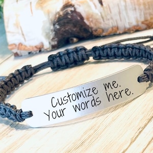 May include: A gray braided cord bracelet with a silver metal tag that says "Customize me. Your words here."