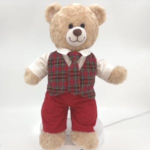 May include: A plush tan teddy bear wearing a white collared shirt, a red and green plaid vest, a matching plaid necktie, and solid red corduroy trousers. The bear has a dark brown stitched nose and a stitched smile on its face.