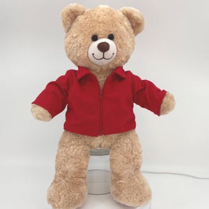 May include: A plush teddy bear with light brown fur, black eyes, and a dark brown nose. The bear is wearing a bright red, long-sleeved, collared jacket that features a vertical zipper closure down the center front. The bear has a stitched smile.