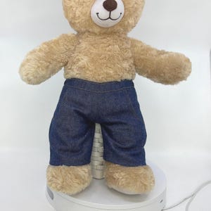 May include: A soft toy teddy bear with light brown fur, dressed in dark blue denim trousers. The bear has a stitched black nose and a smiling mouth. It stands on a white surface.