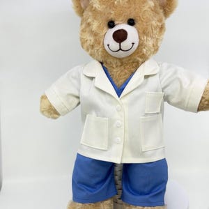 May include: A plush teddy bear wearing a white lab coat over a blue shirt and blue shorts. The bear has a brown nose and black eyes. The lab coat has two pockets and is buttoned up. The bear is standing against a white background.