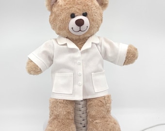 Puede incluir: Un oso de peluche de color marrón claro con ojos oscuros y nariz marrón, que lleva una camisa blanca de manga corta con botones, cuello y dos bolsillos delanteros. El oso tiene un pelaje suave y texturizado y está en posición erguida.