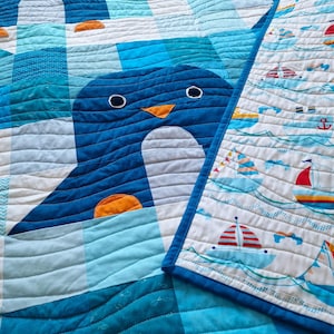 Puede incluir: Un edredón hecho a mano con un gran diseño de pingüino azul y un pico naranja. El edredón tiene un borde de temática náutica con veleros y anclas. El edredón está hecho de tela suave y tiene una textura acolchada.