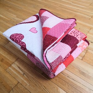 May include: A folded pink and red patchwork quilt with a heart design. The quilt is made of various fabrics with different patterns and textures.