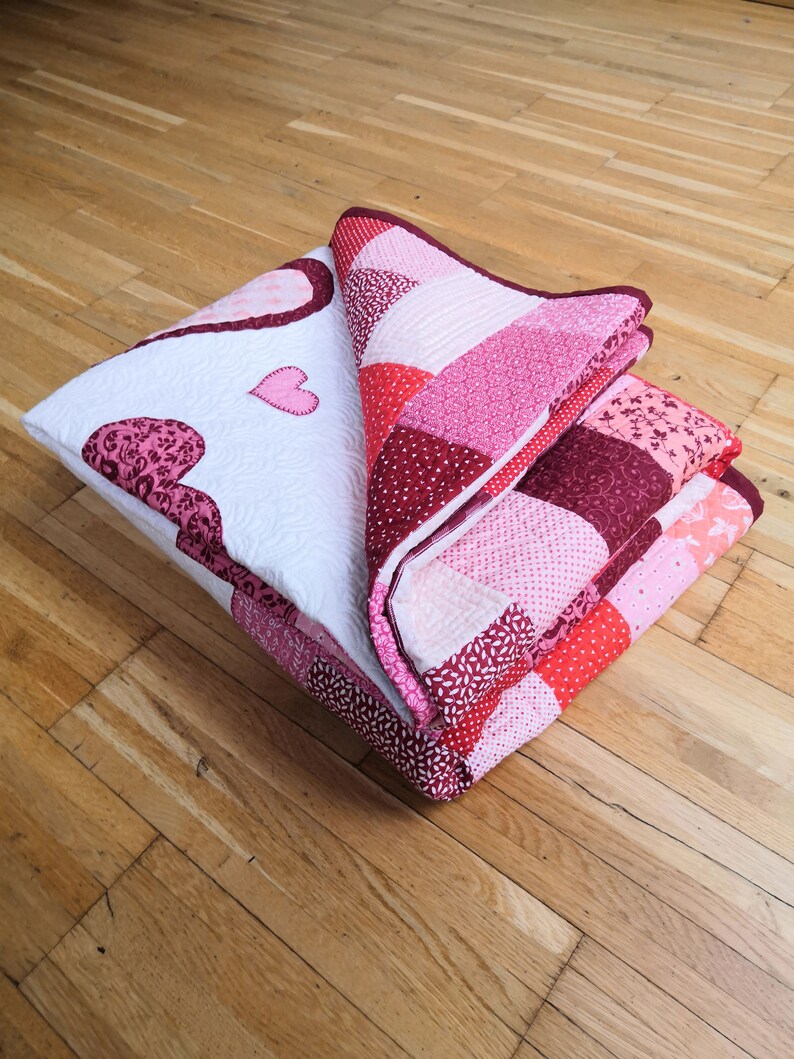 May include: A folded pink and red patchwork quilt with a heart design. The quilt is made of various fabrics with different patterns and textures.