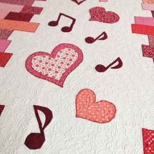 May include: A white quilt with a red, pink, and white patchwork design. The quilt features hearts and musical notes.