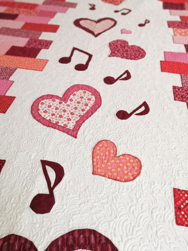 May include: A white quilt with a red, pink, and white patchwork design. The quilt features hearts and musical notes.
