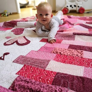 May include: A baby wearing a white and black polka dot shirt lays on a pink and white patchwork quilt. The quilt has a variety of pink and red fabrics with different patterns.