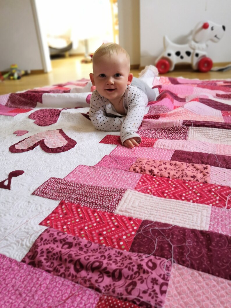 May include: A baby wearing a white and black polka dot shirt lays on a pink and white patchwork quilt. The quilt has a variety of pink and red fabrics with different patterns.
