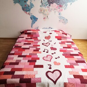 May include: A white quilt with a patchwork design of pink and red squares. The quilt has a row of hearts and musical notes down the center.