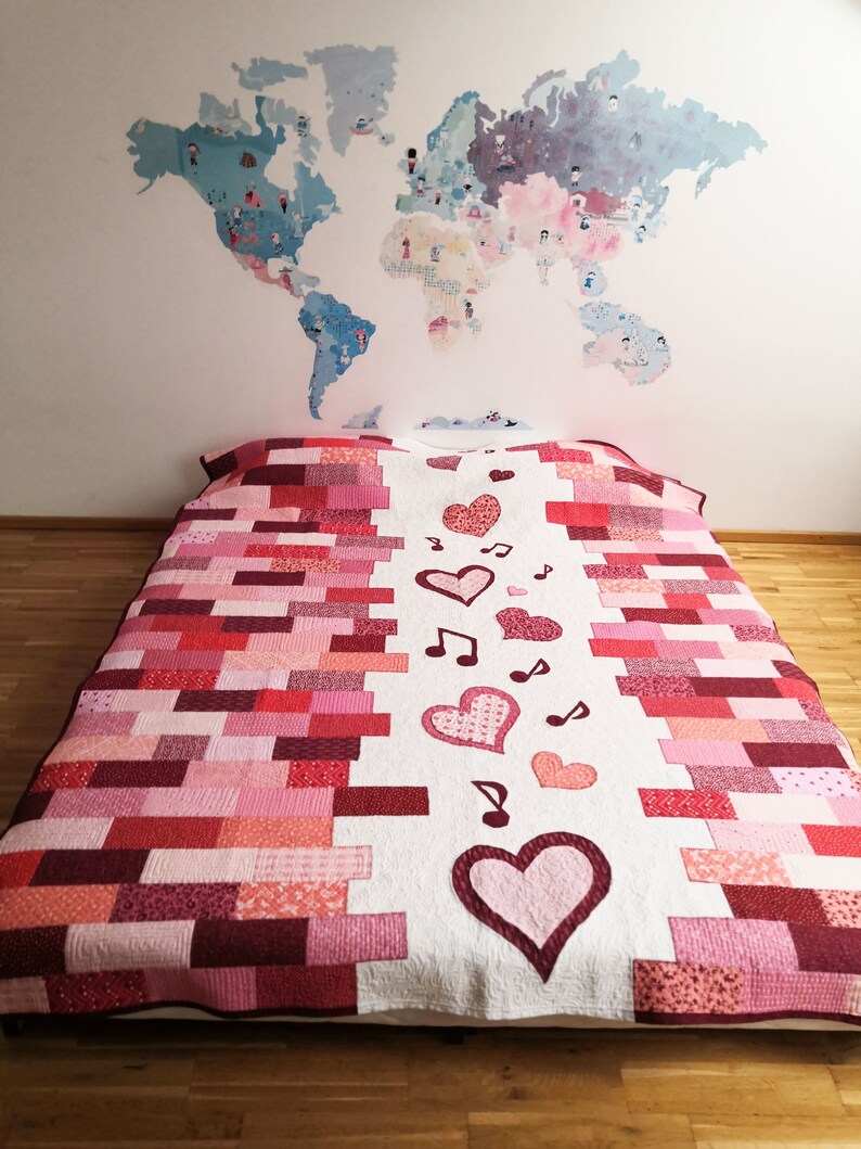 May include: A white quilt with a patchwork design of pink and red squares. The quilt has a row of hearts and musical notes down the center.