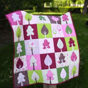 May include: A pink and green patchwork quilt with a tree design. The quilt is made up of squares with different patterns and colors. The quilt is being held up by a person, and the background is a blurry green lawn.