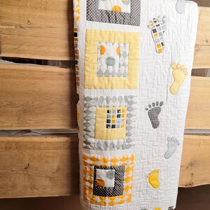 K&ouml;nnte beinhalten: Eine gelbe und graue Babydecke mit wei&szlig;em Hintergrund. Die Decke hat ein Patchwork-Design mit Quadraten und Rauten. Die Decke ist mit Fu&szlig;abdr&uuml;cken verziert.