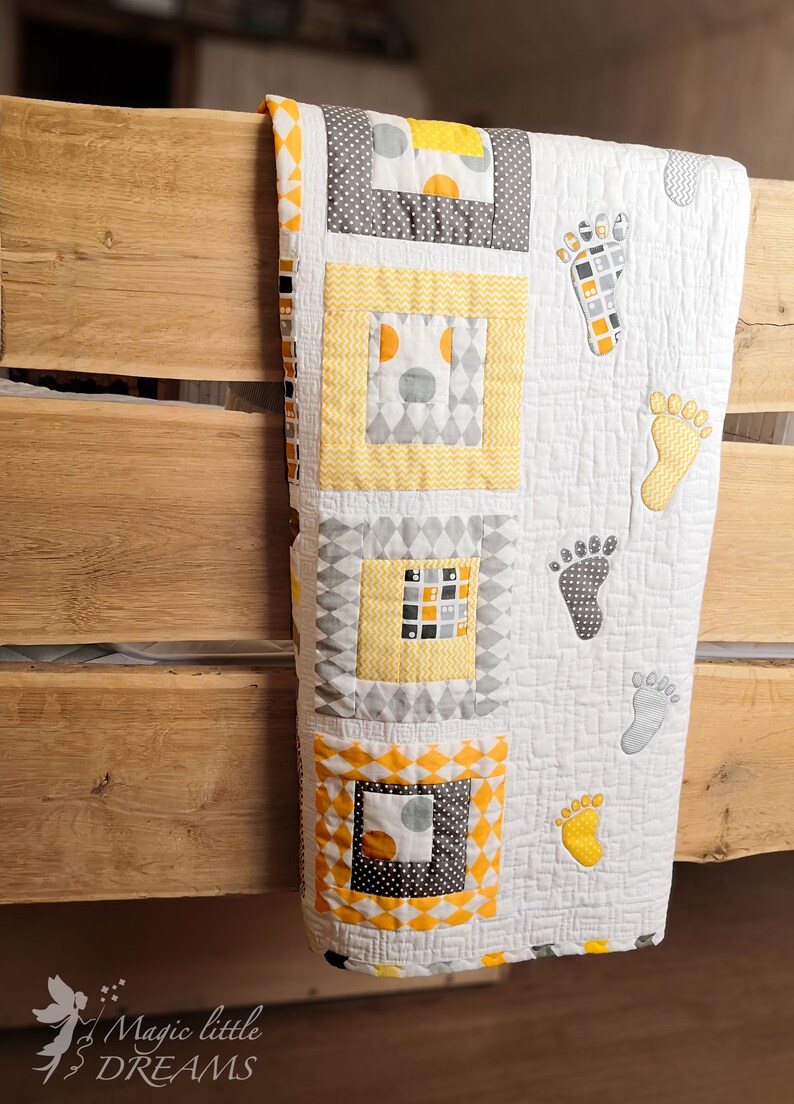 K&ouml;nnte beinhalten: Eine gelbe und graue Babydecke mit wei&szlig;em Hintergrund. Die Decke hat ein Patchwork-Design mit Quadraten und Rauten. Die Decke ist mit Fu&szlig;abdr&uuml;cken verziert.
