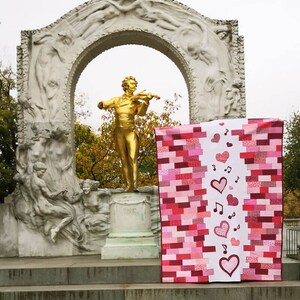 May include: A pink and red patchwork quilt with a white panel featuring hearts and musical notes. The quilt is displayed in front of a white stone archway with a golden statue of a man playing a violin.